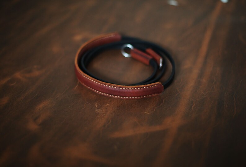 May include: A brown leather camera strap with a black leather lining and a silver metal loop.