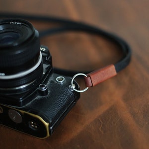 May include: A black camera with a silver lens and a brown leather camera strap. The camera is sitting on a brown wooden surface.