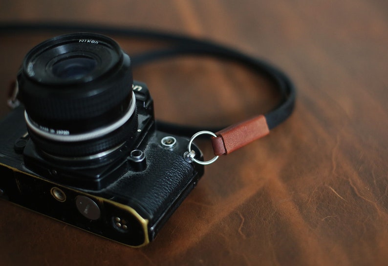 May include: A black camera with a silver lens and a brown leather camera strap. The camera is sitting on a brown wooden surface.