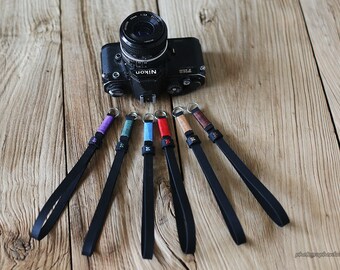 May include: Six camera straps with different colored stitching and metal rings. The straps are black leather and are laid out on a wooden surface.