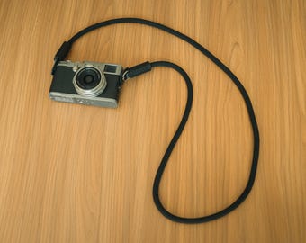 May include: A silver and black digital camera with a circular lens attached to a long, thick black rope strap. The strap features black leather-like connectors at the points where it attaches to the camera body, creating a continuous loop.