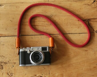 May include: A silver camera with a red and brown camera strap on a wooden surface.