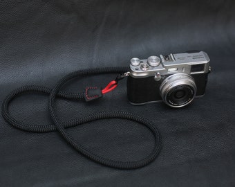 May include: A black camera strap with red stitching, attached to a silver camera with a black lens. The camera is on a black leather surface.