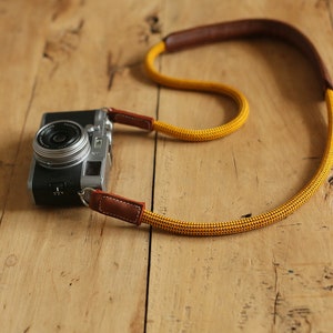 May include: A silver camera with a brown and yellow camera strap. The camera is lying on a wooden surface.