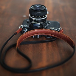 Pode incluir: Uma câmera preta com uma alça de couro marrom. A câmera tem uma lente com um anel prateado. A câmera está em uma superfície de madeira.