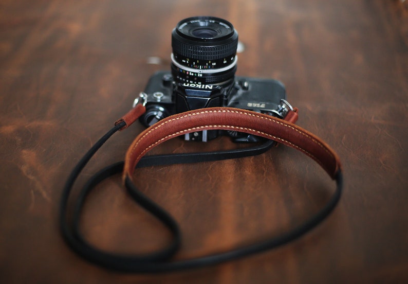May include: A black camera with a brown leather strap. The camera has a lens with a silver ring around it. The camera is sitting on a wooden surface.