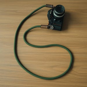 Könnte beinhalten: Eine schwarze Nikon-Kamera mit einem langen, olivgrünen Seil-Kameragurt. Der Gurt verfügt über braune Lederakzente an den Verbindungspunkten, die mit silberfarbenen Metallringen befestigt sind. Das Seil weist eine texturierte, gewebte Oberfläche auf.