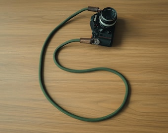 Pode incluir: Uma câmara Nikon preta presa a uma correia de corda comprida de cor verde-azeitona. A correia apresenta detalhes em pele castanha nos pontos de ligação, fixados com anéis de metal prateado. A corda tem uma superfície tecida texturada.