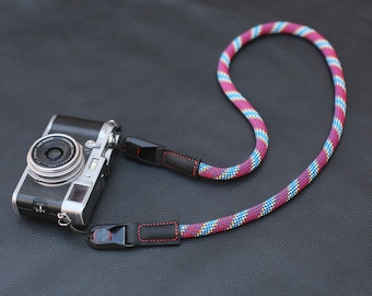 May include: A silver camera with a black leather camera strap with red stitching. The strap is made of a colourful braided rope with red, blue and yellow stripes.