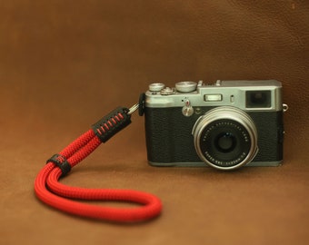 May include: A silver camera with a black leather strap and a red rope wrist strap. The camera has a black lens with the text "FUJIFILM SUPER EBC 142mm 1:2.8" printed on it.