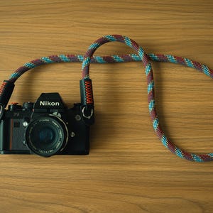 May include: A black Nikon camera with a lens attached, featuring a long, thick, braided rope camera strap. The strap has a repeating pattern of teal, red, and dark blue colors, with black leather connectors secured by bright orange stitching.