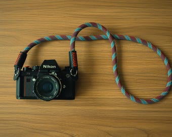 Pode incluir: Uma câmara Nikon preta com lente, apresentando uma correia de corda entrançada longa e grossa. A correia tem um padrão repetitivo de cores azul-petróleo, vermelho e azul escuro, com conectores de couro preto fixados por costuras laranja.