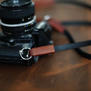 May include: A black camera with a brown leather strap. The camera has a silver ring around the lens and a brown leather strap with a silver ring.