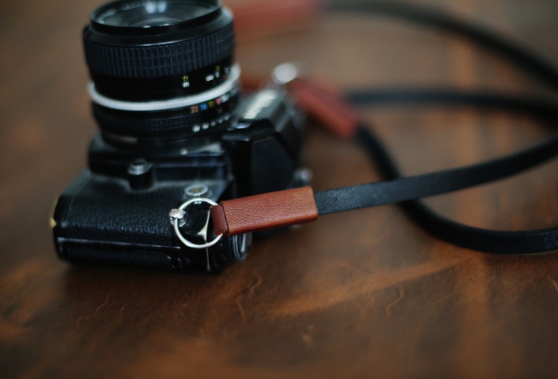 May include: A black camera with a brown leather strap. The camera has a silver ring around the lens and a brown leather strap with a silver ring.