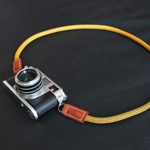 May include: A vintage camera with a silver lens and black leather body, attached to a yellow braided camera strap with brown leather ends. The strap is approximately 40 inches in length.