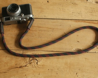 Op de afbeelding: Een zwarte en rode touwen camerariem bevestigd aan een zilveren en zwarte camera. De riem is gemaakt van een dik, duurzaam touw met rode accenten. De camera is een compact model in retrostijl.