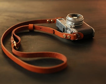 May include: A brown leather camera strap with gold metal accents and a silver camera attached.