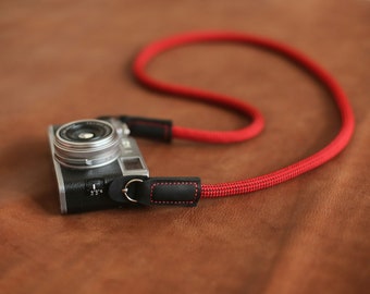 Op de afbeelding: Een zilveren camera met een rode en zwarte cameraband. De band is gemaakt van dik, gevlochten touwmateriaal.