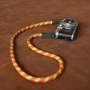Correa para cámara con diseño naranja: cuerda de escalada hecha a mano con detalles dorados y extremos de cuero.