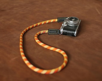 May include: A camera with a black and silver body and a silver lens is attached to a bright orange and yellow camera strap.