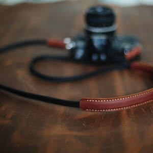 May include: A brown leather camera strap with a black strap and white stitching. The strap is lying on a wooden surface.