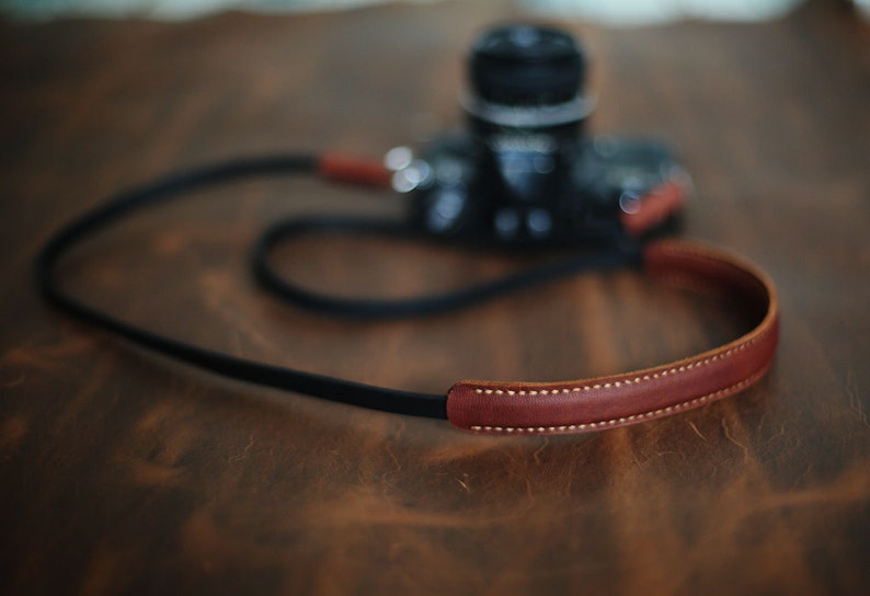May include: A brown leather camera strap with a black strap and white stitching. The strap is lying on a wooden surface.