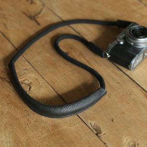 May include: A black camera strap with a textured surface. The strap is made of leather and has a simple design.