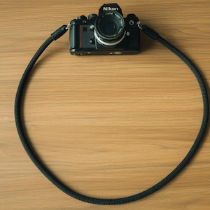 Könnte beinhalten: Eine schwarze Nikon-Kamera mit einem runden, schwarzen Kameragurt aus Seil, der an den seitlichen Ösen befestigt ist. Der Gurt ist in einem großen Kreis angeordnet und zeigt seine texturierte, geflochtene Kordelstruktur und schwarze Lederverbinder.