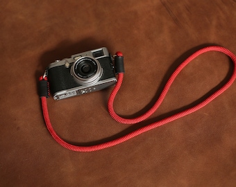 May include: A silver camera with a red camera strap on a brown leather surface.