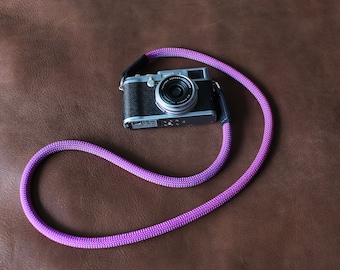 May include: A purple camera strap with a silver camera attached to it. The camera strap is made of a braided material and has a textured look. The camera is a compact model with a black body and a silver lens.