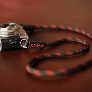 May include: A silver camera with a black leather strap attached. The strap is made of black and red rope with a red stitching detail.