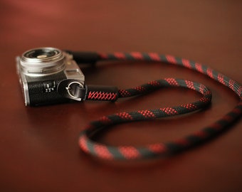 Op de afbeelding: Een zilveren camera met een zwarte leren band. De band is gemaakt van zwart en rood touw met een rood stikseldetail.