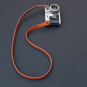 May include: A silver and black camera with a brown leather strap. The camera lens is facing forward. The strap is thin and appears to be made of leather. The camera and strap are on a dark surface.