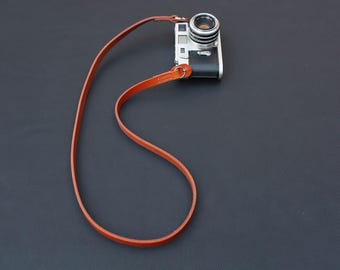 May include: A silver and black camera with a brown leather strap. The camera lens is facing forward. The strap is thin and appears to be made of leather. The camera and strap are on a dark surface.