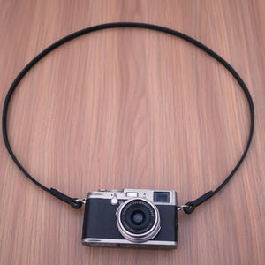 May include: A silver and black digital camera with a circular lens and a thin black neck strap attached to both sides. The camera body features a metallic silver top plate and a textured black grip, with a centred lens and a viewfinder window.