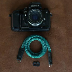 Peut inclure: Appareil photo Nikon F4 noir avec objectif, une sangle de caméra turquoise et deux anneaux en caoutchouc noirs sur une surface marron. La sangle est en corde turquoise avec des extrémités en cuir noir et des anneaux métalliques.