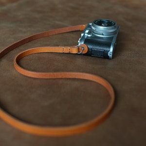 Può includere: Un cinturino per fotocamera in pelle marrone con un anello argentato e una fotocamera argentata attaccata.