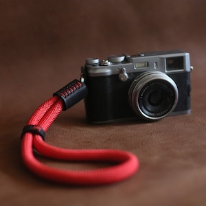 May include: A silver camera with a black lens and a red camera strap. The camera is sitting on a brown surface.