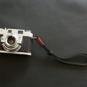 Può includere: Una fotocamera vintage argento e nera con un cinturino da polso marrone e nero. La fotocamera ha un obiettivo prominente e vari quadranti e pulsanti. Il cinturino è attaccato al lato della fotocamera.