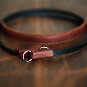May include: A brown leather camera strap with a black leather strap attached to it. The strap has two silver metal rings.