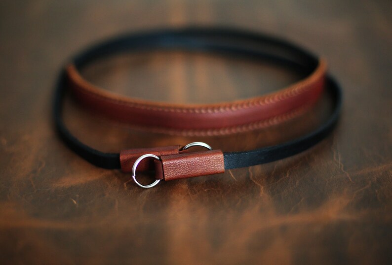 May include: A brown leather camera strap with a black leather strap attached to it. The strap has two silver metal rings.