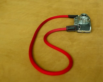 May include: A silver camera with a bright red neck strap. The strap is made of a thick, woven material and has black leather accents where it attaches to the camera. The camera is resting on a tan surface.