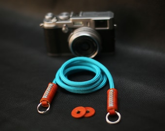 May include: A turquoise camera strap with orange leather accents and silver rings. The strap is made of a braided rope material.