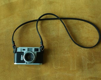 May include: A black and silver camera with a black strap lies on a textured, golden-yellow surface. The camera has a prominent lens and a sleek, minimalist design, with the strap looped above.