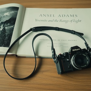 Op de afbeelding: Een zwarte Nikon filmcamera met een lange, dunne zwarte leren riem die op een open fotoboek ligt met de titel Ansel Adams Yosemite and the Range of Light. De camera is voorzien van een ronde lens en metalen onderdelen voor de bevestiging.