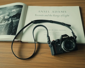 May include: A black Nikon film camera with a long, thin black leather strap resting on an open photography book titled Ansel Adams Yosemite and the Range of Light. The camera features a circular lens, a silver shutter button, and metal hardware.