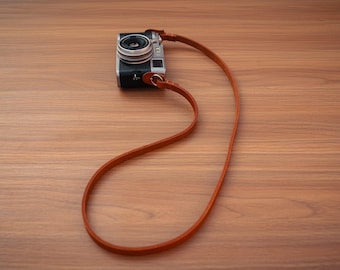 May include: A compact digital camera with a silver and black body, featuring a lens on the front and a long, thin, tan leather strap attached to the sides, forming a large loop that extends outward from the camera body.