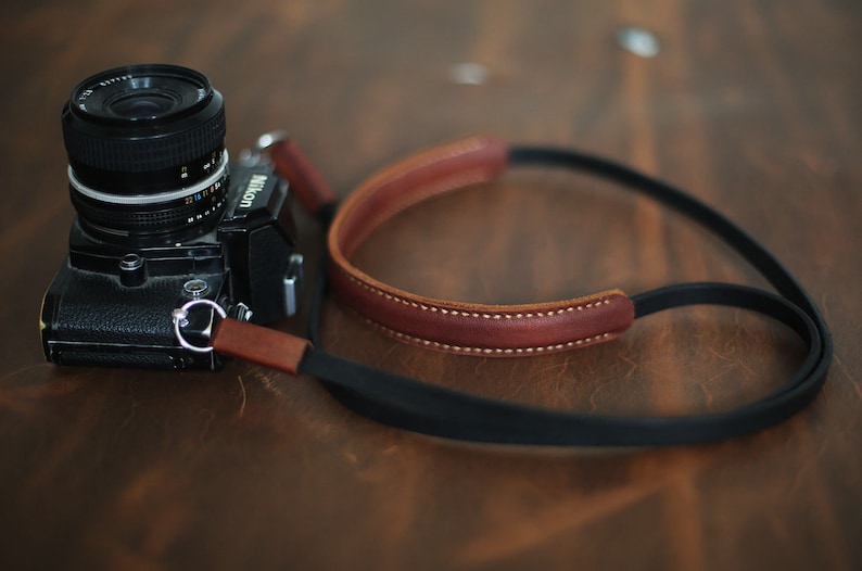 May include: A black Nikon camera with a brown leather camera strap. The camera has a silver ring around the lens.