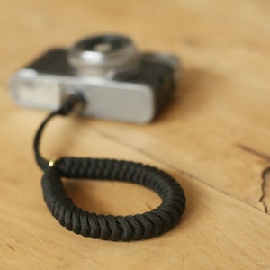 Peut inclure: Un sangle de caméra noire en cordon tressé avec une boucle en métal doré. La sangle est attachée à un appareil photo argenté.
