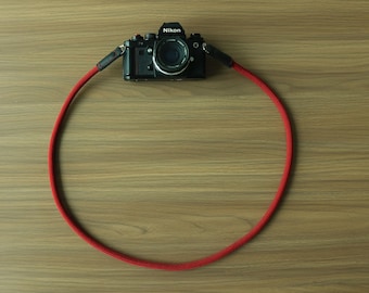 May include: A black Nikon camera attached to a long, circular red rope camera strap. The strap is made of a woven, textured material and features dark leather connectors at the points where it attaches to the camera body on both sides.